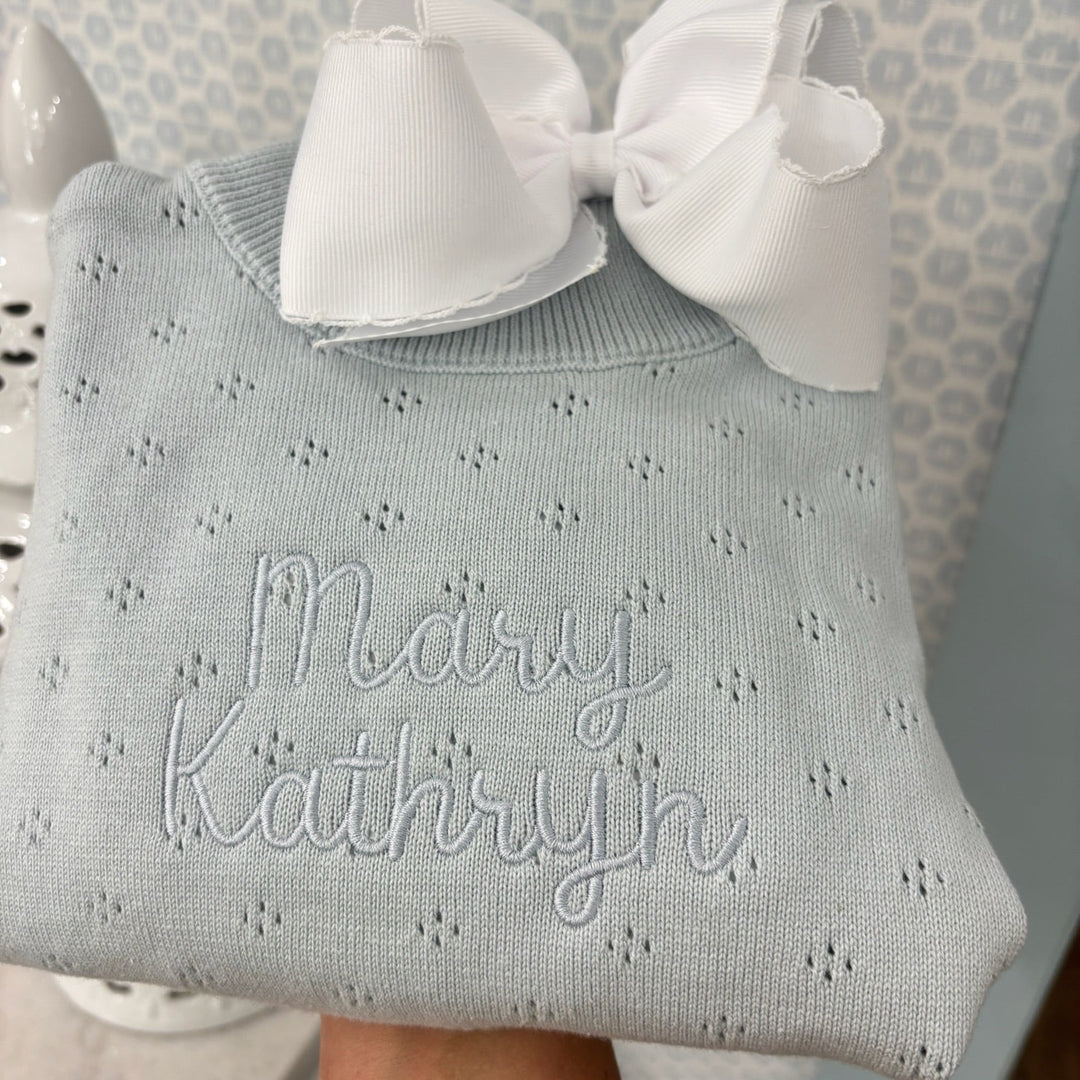 Light blue baby outfit with white bow and embroidered name against a neutral background