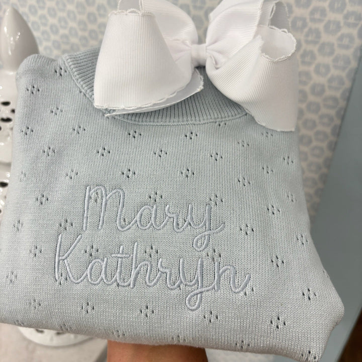 Light blue baby outfit with white bow and embroidered name against a neutral background